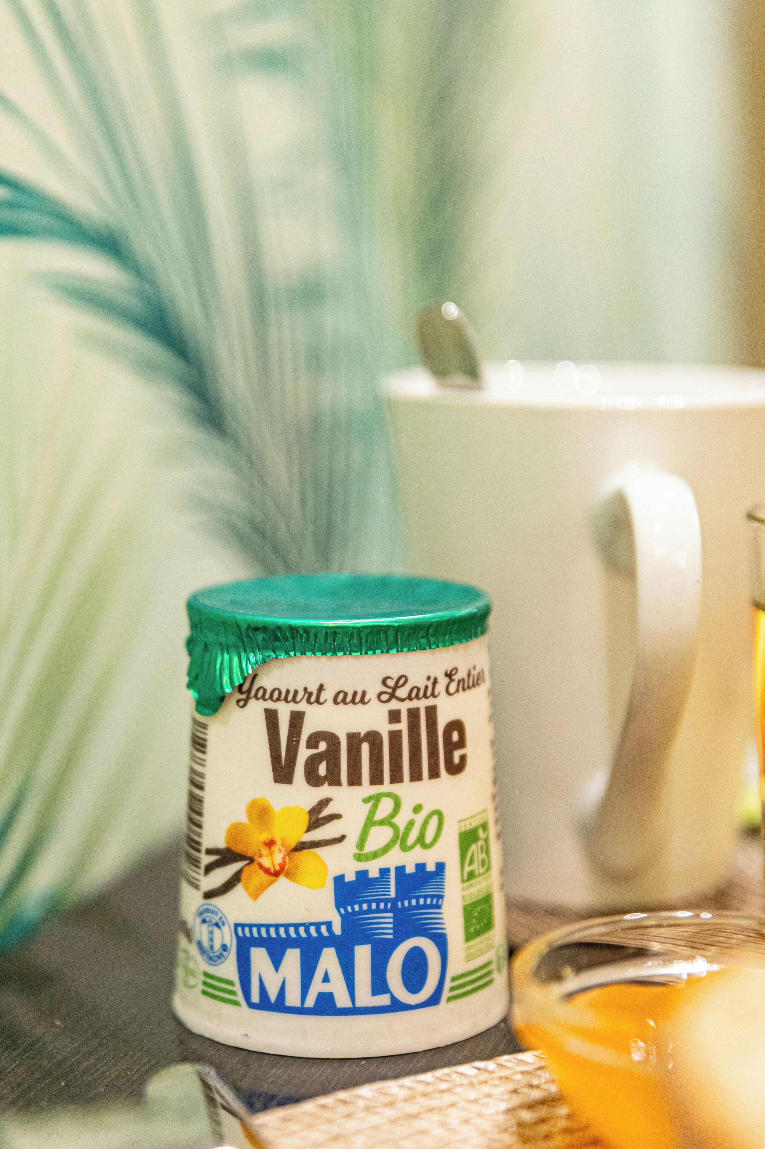 Un pot de yaourt nature bio à la vanille, une tasse blanche et un verre contenant un liquide jaune.
