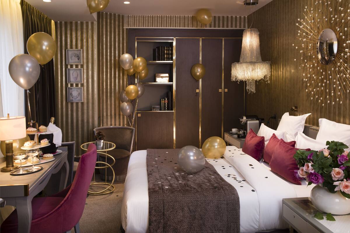 A hotel room with a bed, bedside table, lamp, chair, and a desk with a lamp and a small plant.