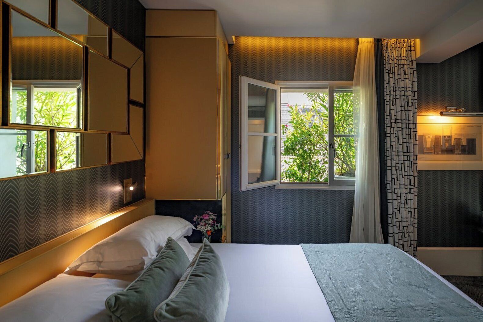 A hotel room with a bed, a window with a view of greenery, a mirror, and a wall with patterned wallpaper.