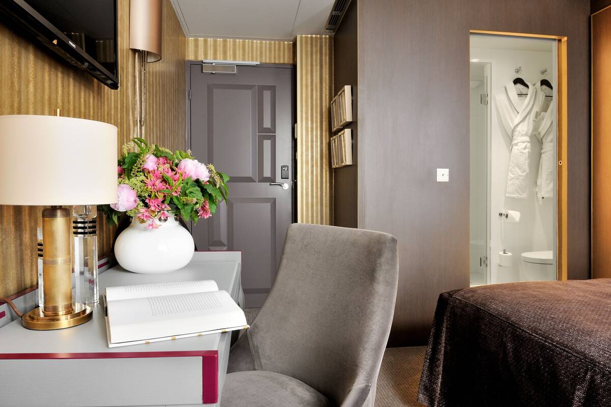 A hotel room with a desk, a chair, a bed, a lamp, a vase with flowers, and a wardrobe with clothes.