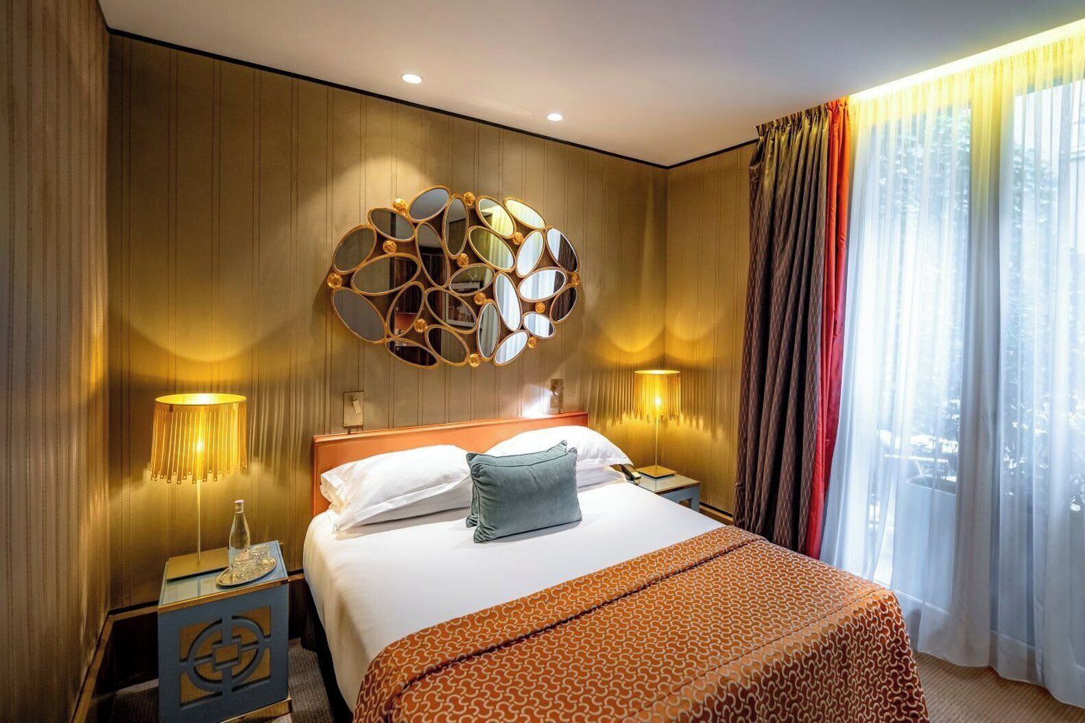 Una camera d'hotel con un letto grande, uno specchio decorativo, due lampade da comodino e vista all'esterno tramite tende leggere.
