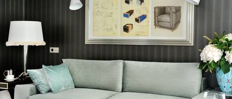 A modern living room with a grey sofa, a blue side table, a white lamp, and a framed picture of furniture designs on the wall.