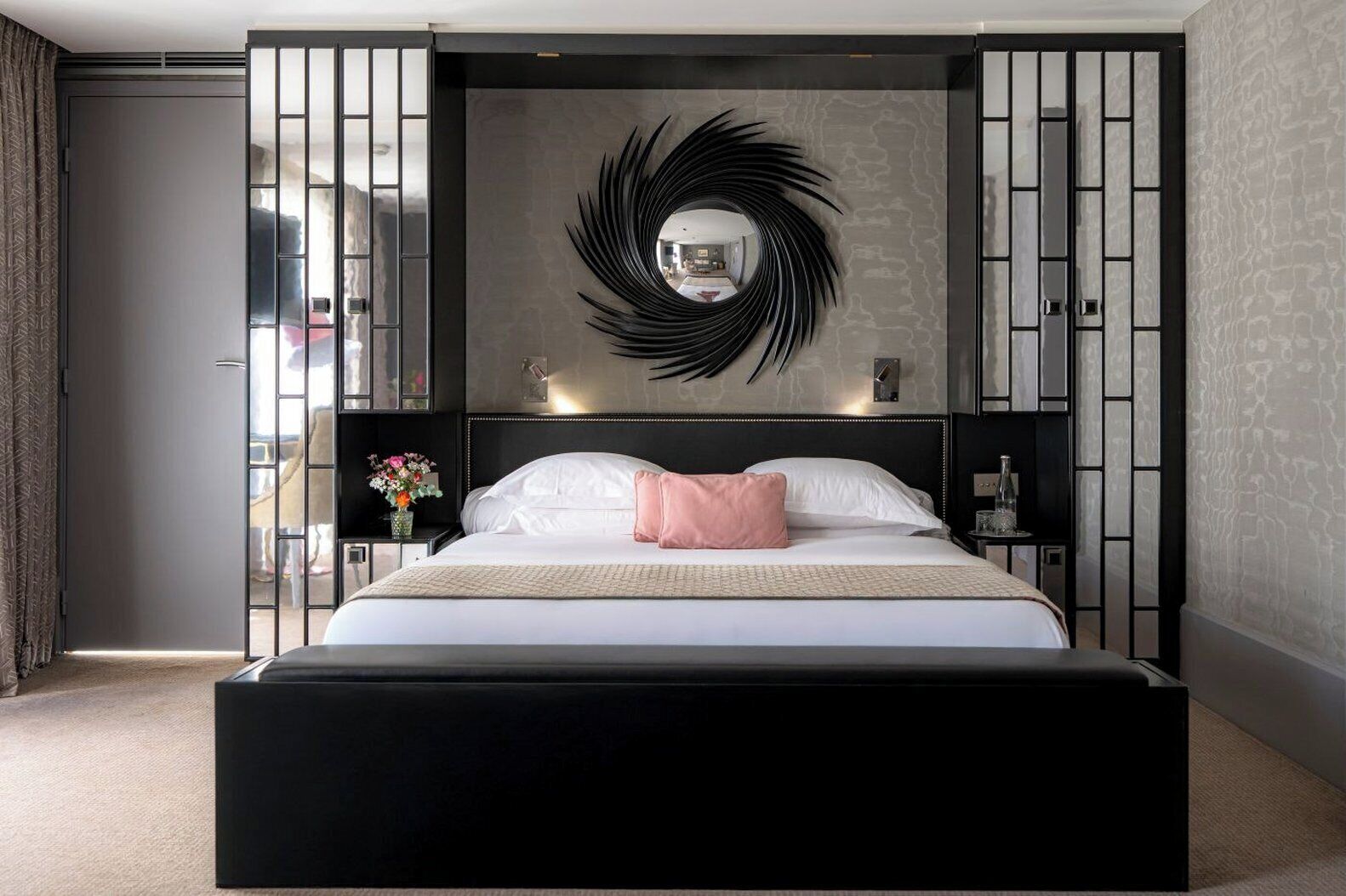 A modern hotel room with a large bed, a decorative wall mirror, and a view of the outdoors.