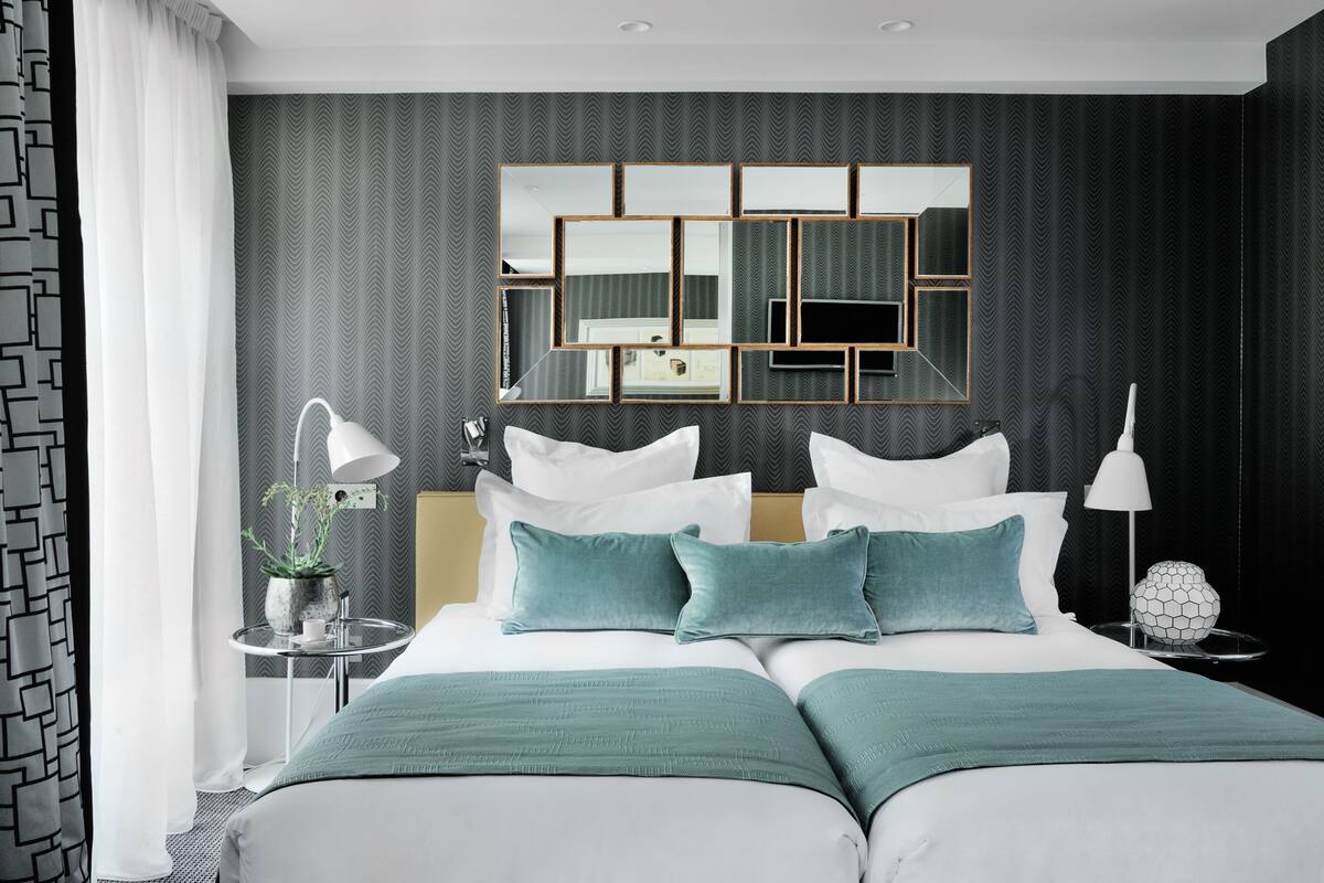 A modern hotel room with a large bed, a mirror with geometric frames, a bedside table with a lamp, and a decorative sphere.