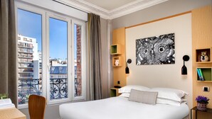 Habitación de hotel con una cama grande, un escritorio, una silla, un balcón con vistas a edificios y un cuadro en la pared.