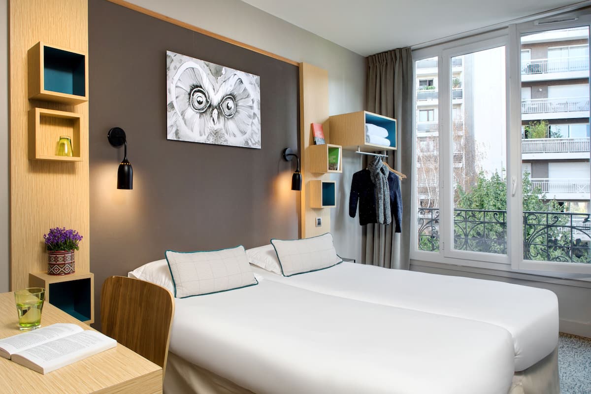Een hotelkamer met een groot bed, een bureau met stoel, een raam met uitzicht op gebouwen en een wandplank met een bloevas.
