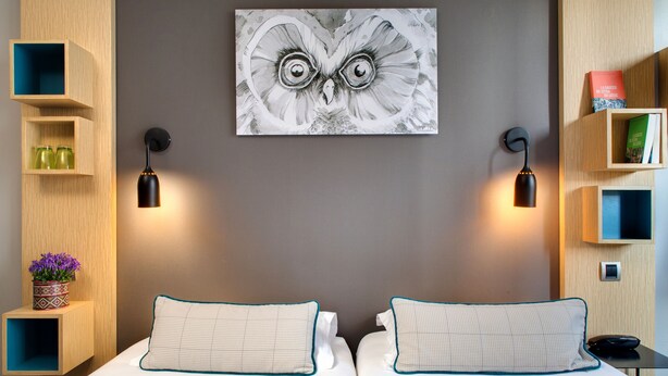 Habitación de hotel con dos camas, un cuadro colgado en la pared de un búho y estanterías empotradas con libros y objetos decorativos.