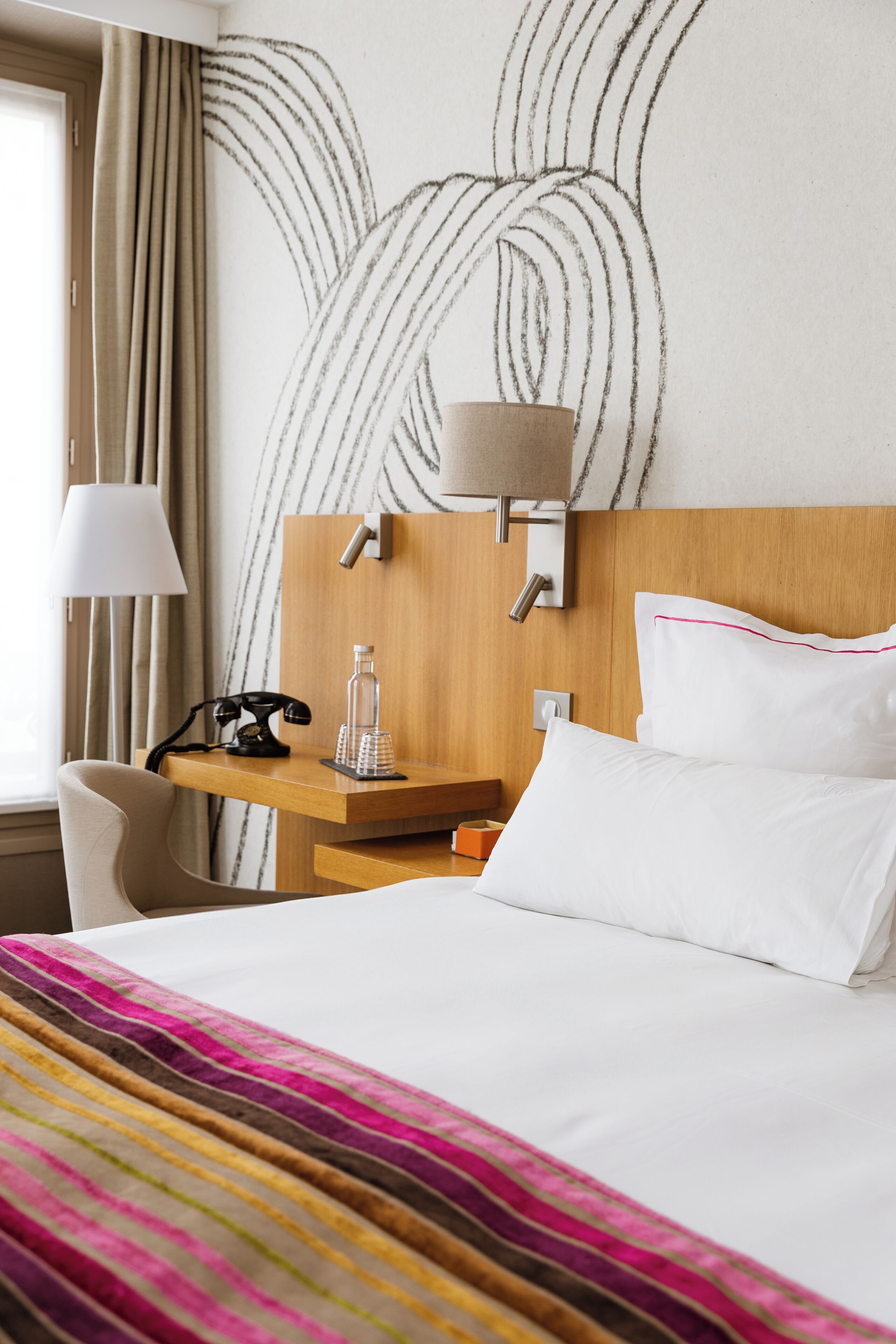 A hotel room with a bed, a desk with a lamp, a telephone, and a wall with a leaf pattern.
