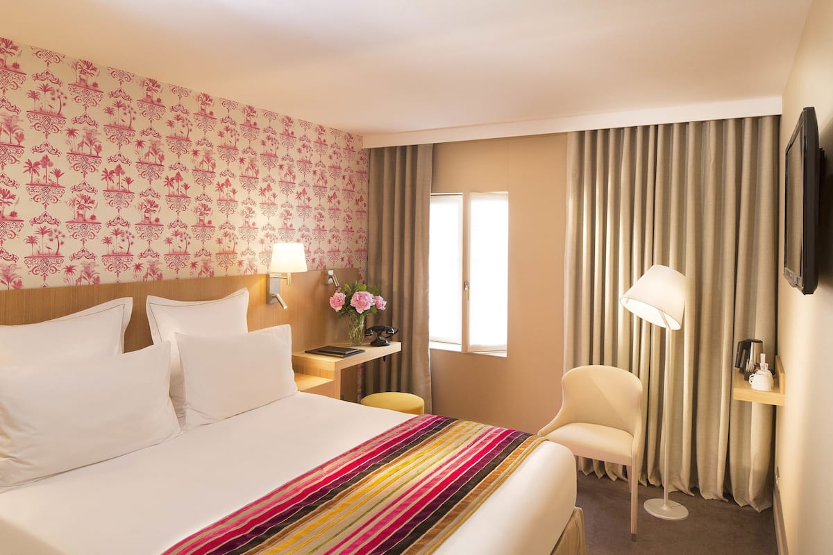 A hotel room with a large bed, a desk with a lamp, a chair, and a TV on the wall.