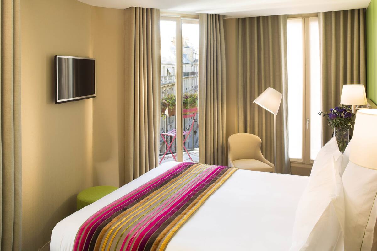 A hotel room with a bed, a flat-screen TV mounted on the wall, a chair, a small stool, and a window with curtains.