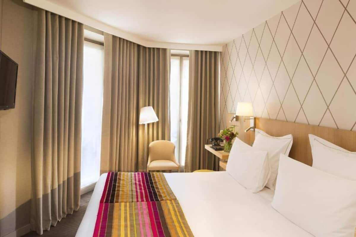 A hotel room with a large bed, a desk with a lamp, a chair, and a view of the outside through curtains.