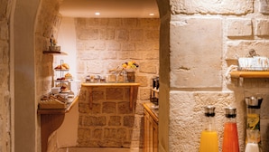 Un couloir voûté en pierre mène à un comptoir où l’on prend le petit-déjeuner. Les murs sont en pierre, le sol est carrelé et une étagère contient des boissons.