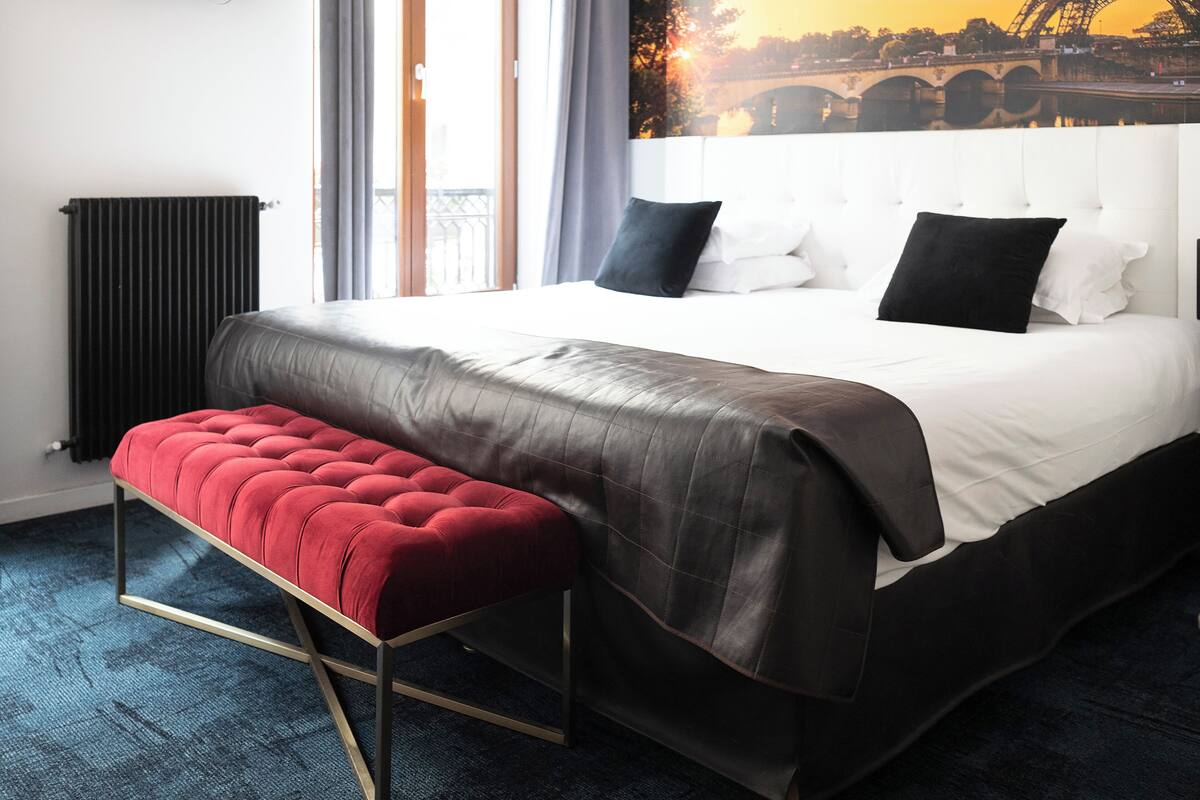 A hotel room with a large bed, a red bench, a radiator, and a view of a bridge and sunset.