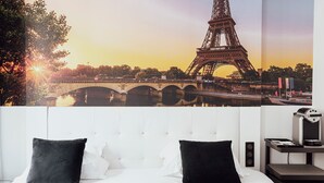 A hotel room with a large poster of the Eiffel Tower, a bed with white and black pillows, and a bedside table with a lamp and a small speaker. - Hotel Aida Marais Printania (Paris)