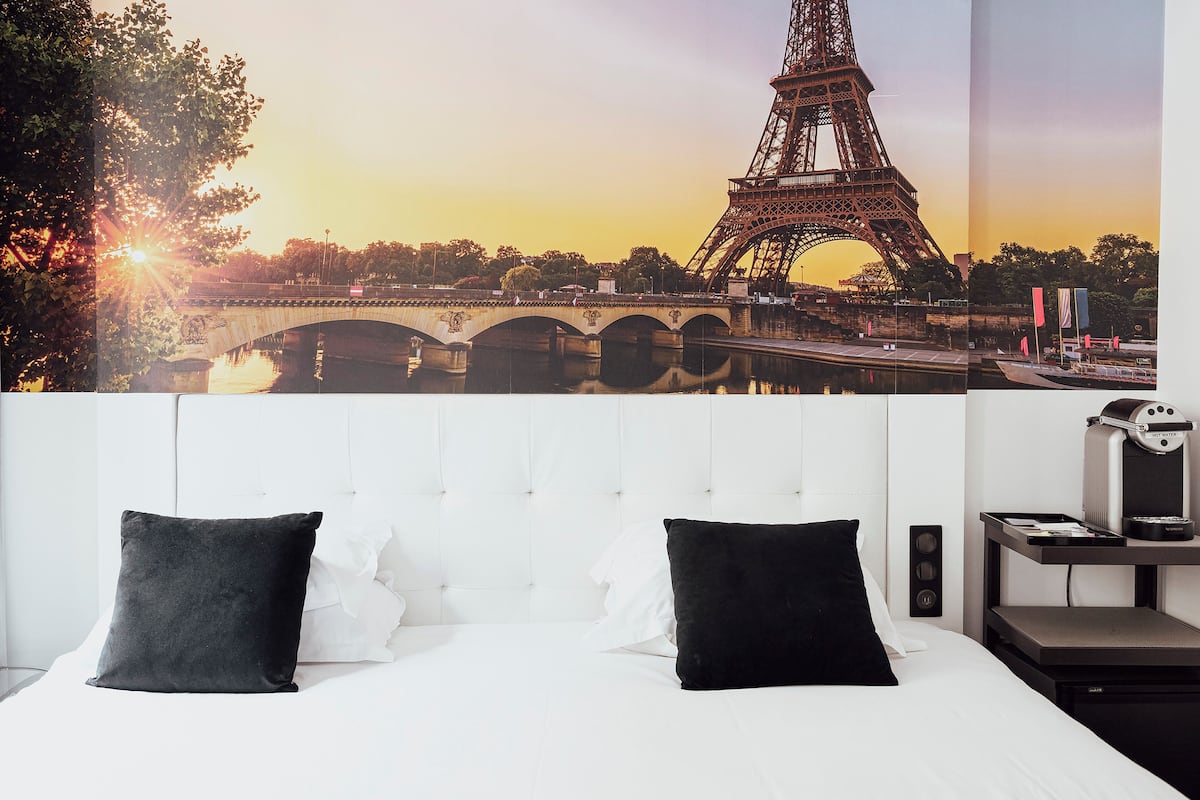 A hotel room with a large poster of the Eiffel Tower, a bed with white and black pillows, and a bedside table with a lamp and a small speaker.