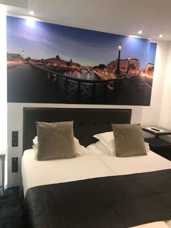 A hotel room with a bed, two pillows, a nightstand, and a wall-mounted picture of a bridge. - Hotel Aida Marais Printania (Paris)