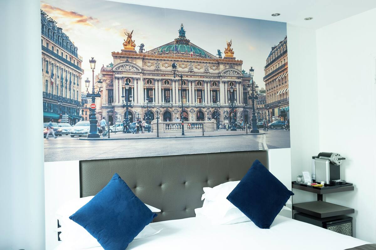A hotel room with a large bed, two blue pillows, a headboard, a nightstand with a coffee maker, and a view of a historic building.