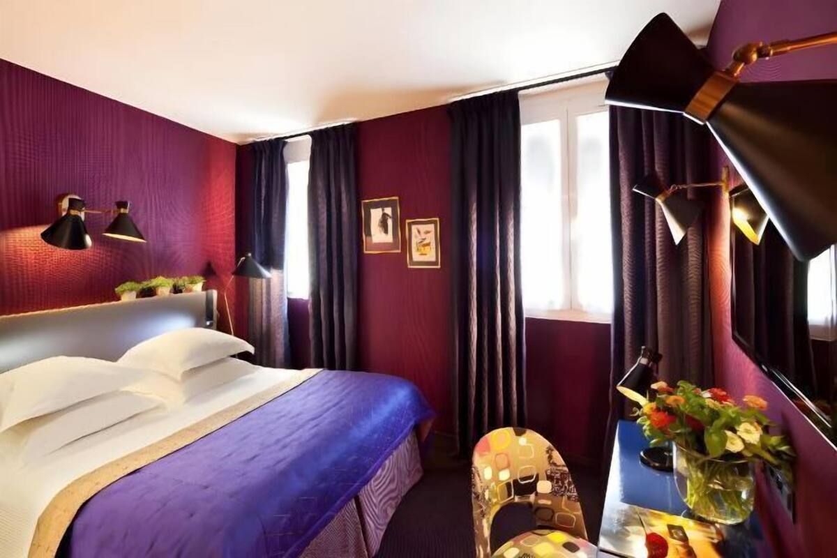 A hotel room with a bed, bedside lamps, a small table with a vase of flowers, and a window with curtains.