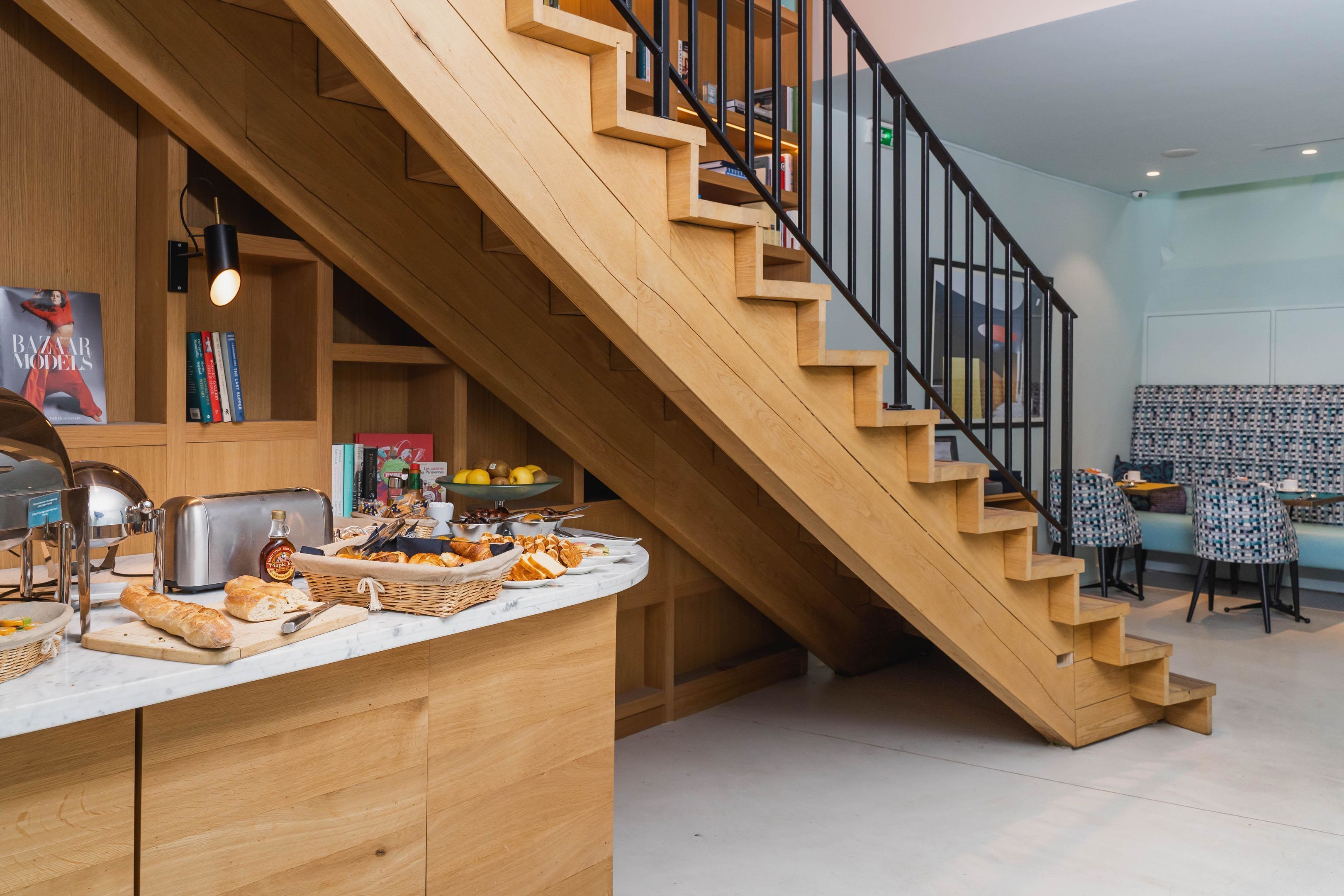 Une cuisine moderne avec un escalier en bois, un comptoir-buffet où sont disposés du pain et des pâtisseries, et un coin repas avec des chaises à motifs.