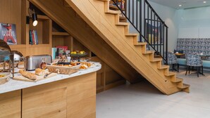 A modern kitchen with a wooden staircase, a buffet counter with bread and pastries, and a dining area with patterned chairs.