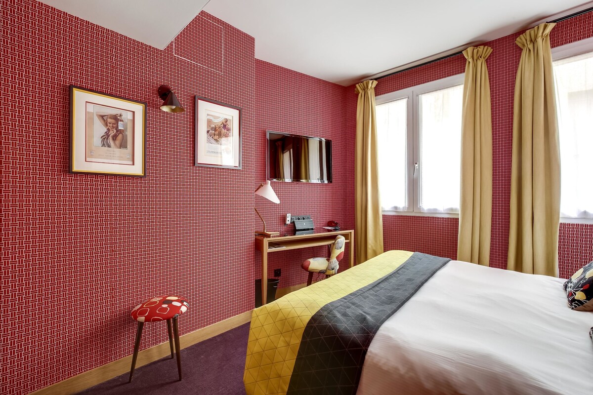A hotel room with a red patterned wall, a bed with a quilt, a desk with a chair, a mirror, and a window with curtains.