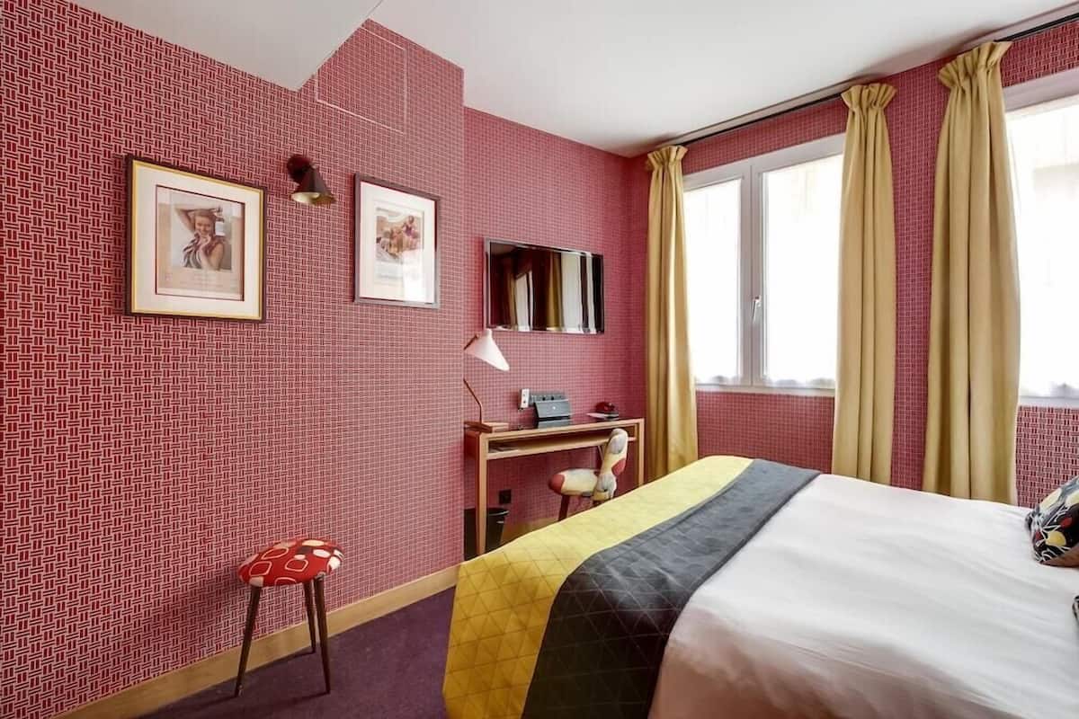 A hotel room with a patterned red wall, a bed with a black and yellow bedspread, a desk with a lamp, a chair, and a window with curtains.