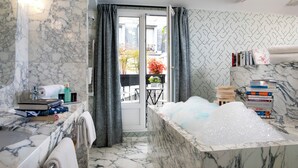 A bathroom with a marble sink, a bathtub filled with bubbles, and a view of the outside through a door. - Hôtel Artus (Paris)