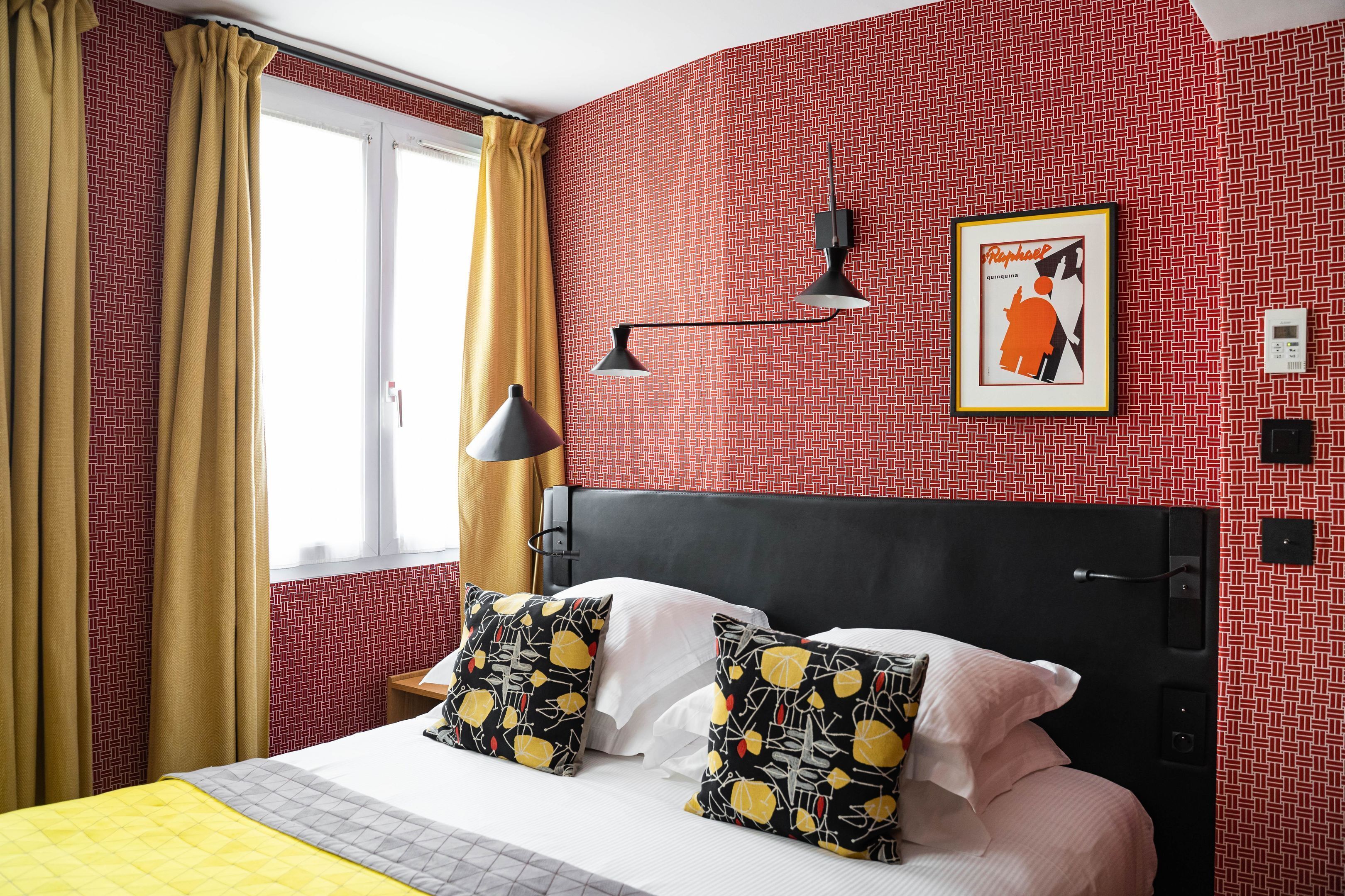 A hotel room with a patterned red wall, a bed with white and yellow bedding, a black headboard, and a framed picture on the wall.