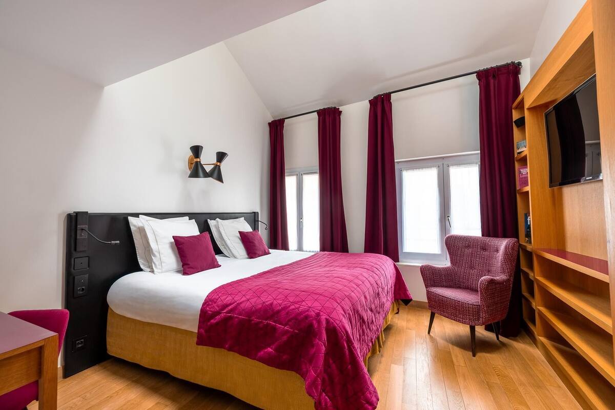 A modern hotel room with a large bed, a desk, a chair, a TV, and a window with curtains.