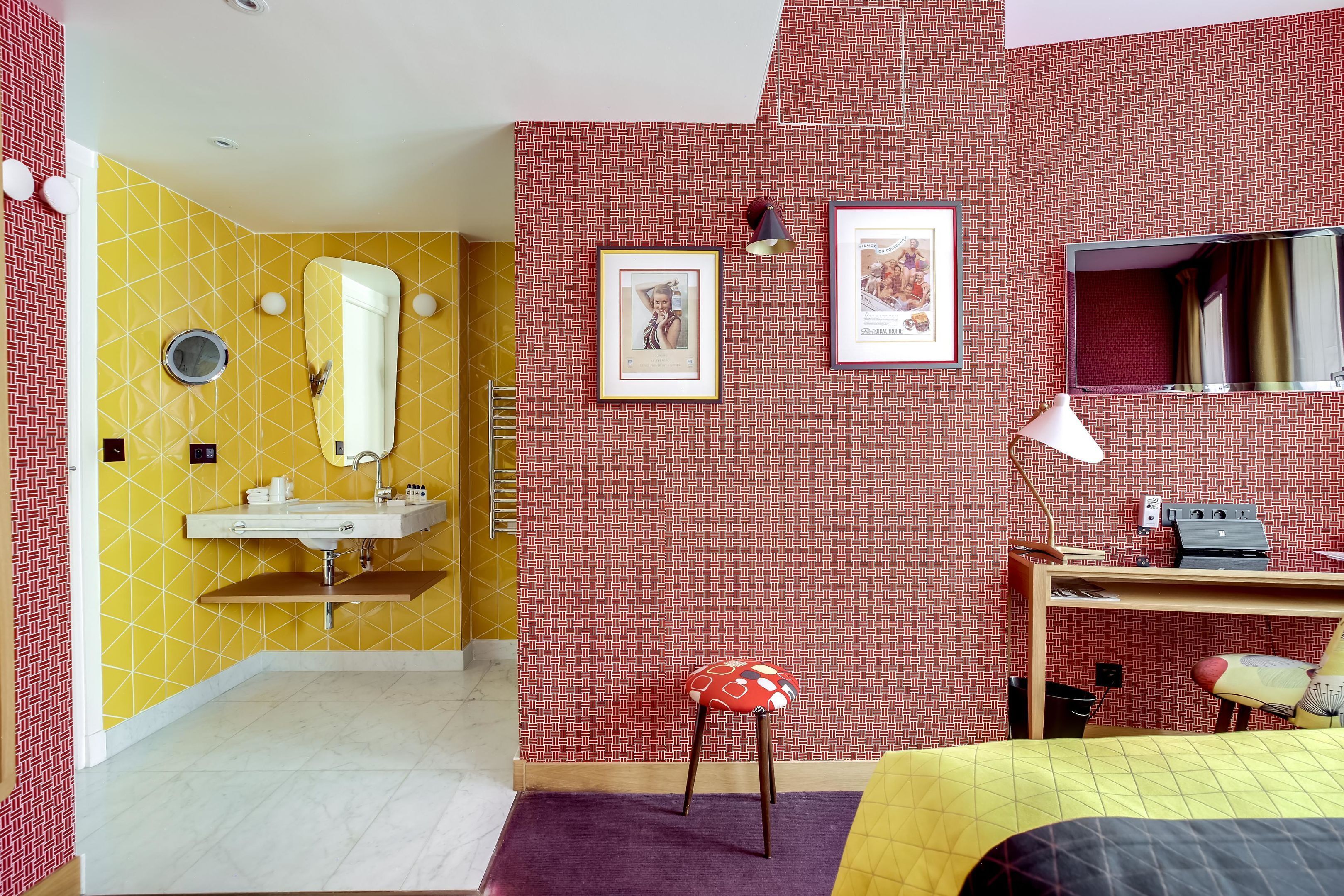 A hotel room with a yellow and red patterned wall, a bed with a yellow and black bedspread, a desk with a chair, a mirror, and a small stool.
