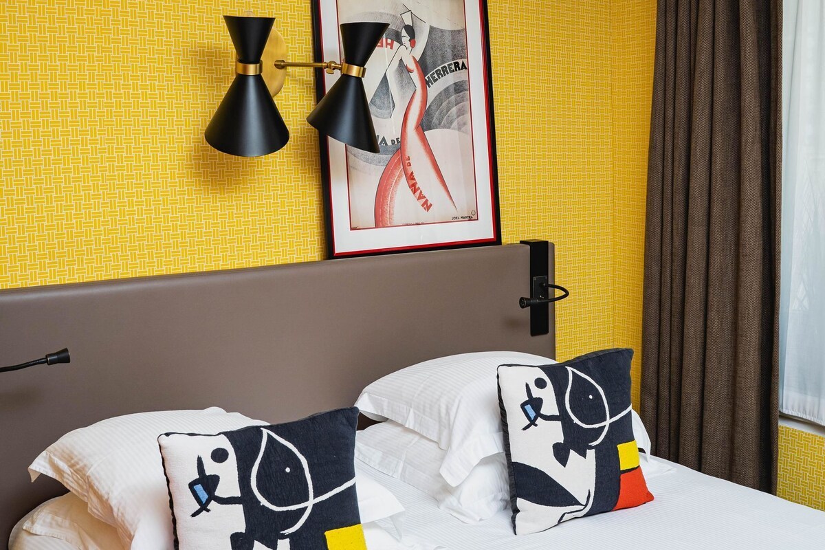 A hotel room with a bed, two pillows with a skull design, a framed picture on the wall, and a black lamp.