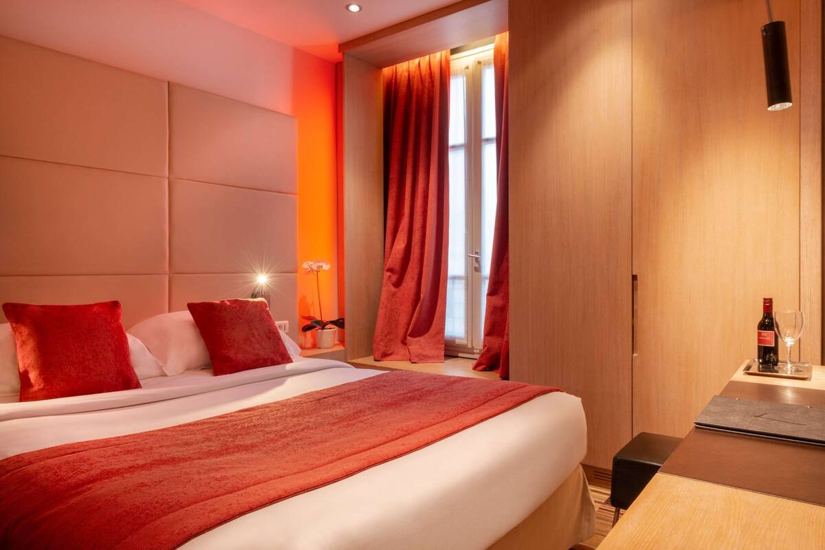 A hotel room with a large bed, red bedding, a wooden desk with a wine bottle, and a window with curtains.