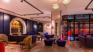 A modern lounge area with sofas, armchairs, and a coffee table. There is a bookshelf and a large window with a view of the outdoors. - Golden Tulip Opera De Noailles (Paris)