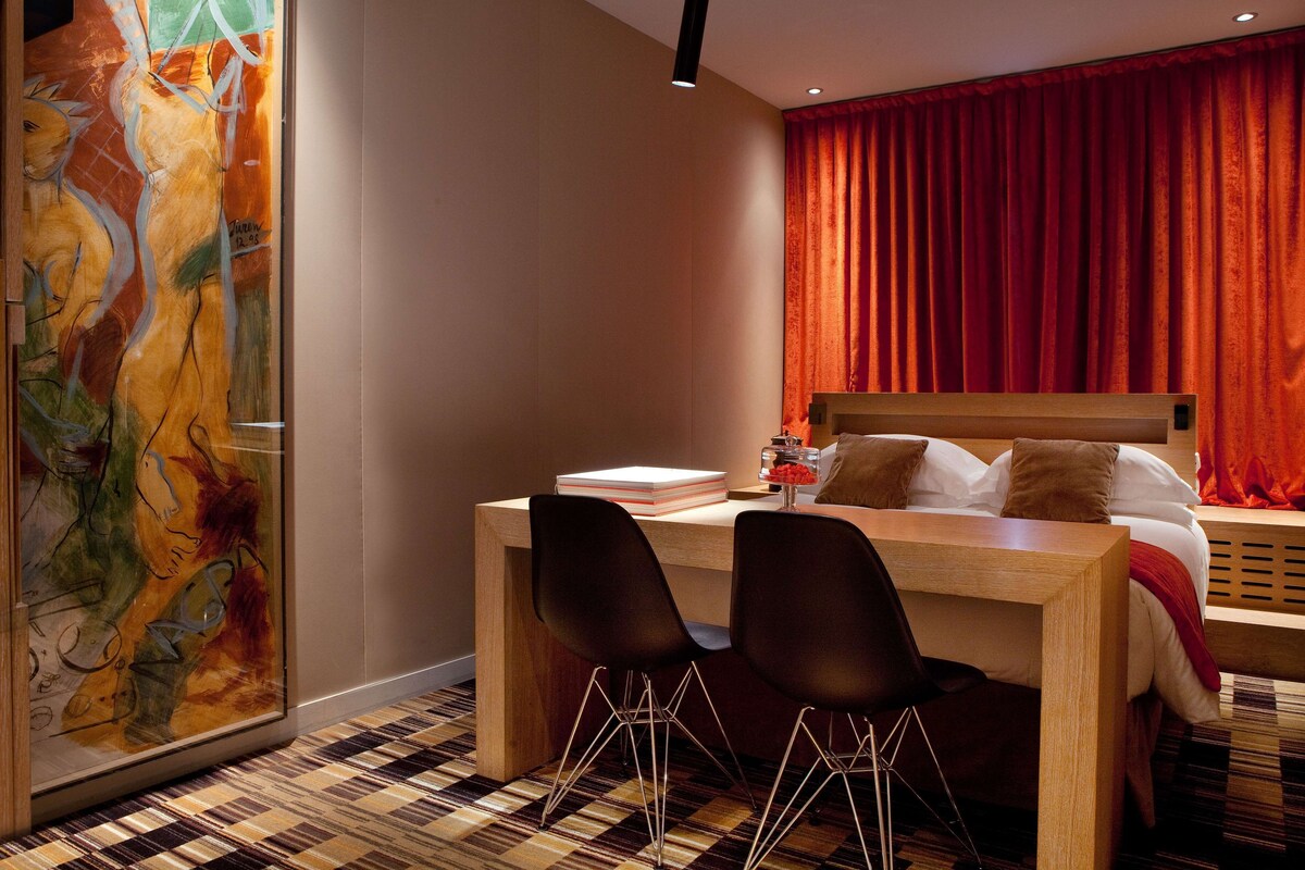 Habitación de hotel con cama, escritorio con sillas, un cuadro abstracto grande en la pared y una cortina roja.