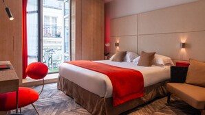 A hotel room with a large bed, a red chair, a sofa, and a view of the city through a large window. - Golden Tulip Opera De Noailles (Paris)