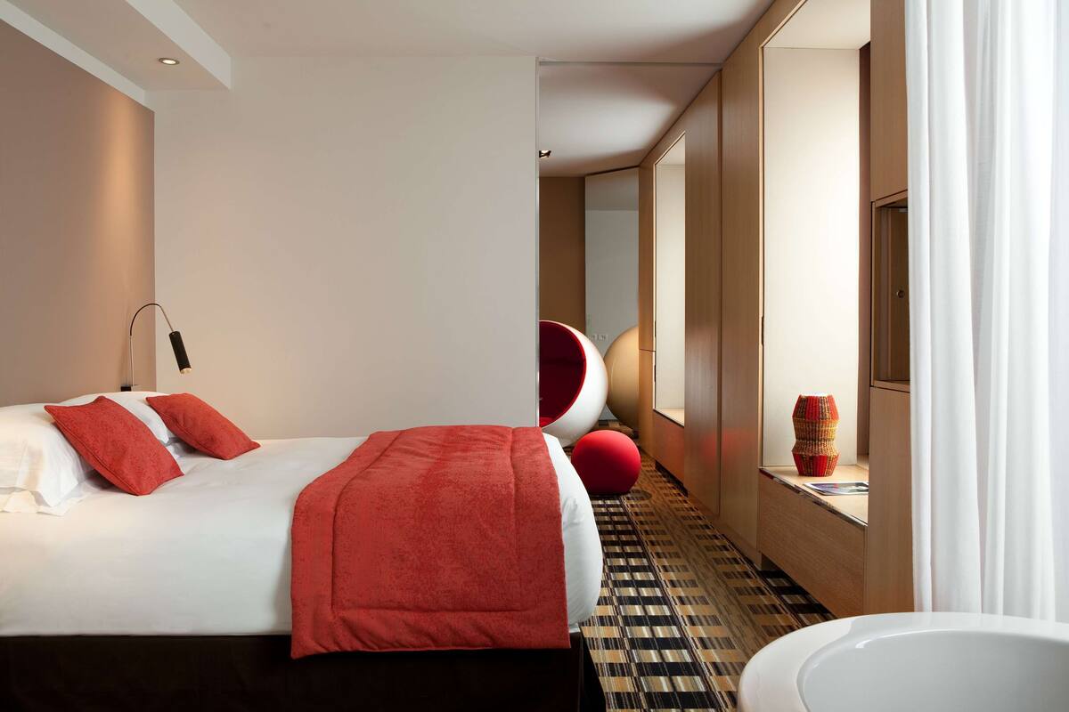 A modern hotel room with a large bed, a red accent wall, a round mirror, and a bathroom with a bathtub.