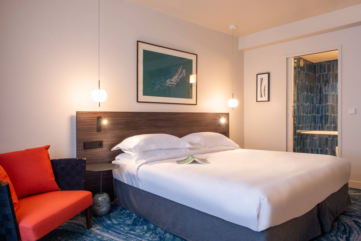 A hotel room with a large bed, a red chair, a framed artwork on the wall, and a glass door leading to a bathroom.
