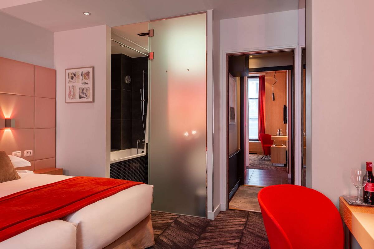 A hotel room with a bed, a red chair, a desk with a wine glass, and a bathroom visible through an open door.