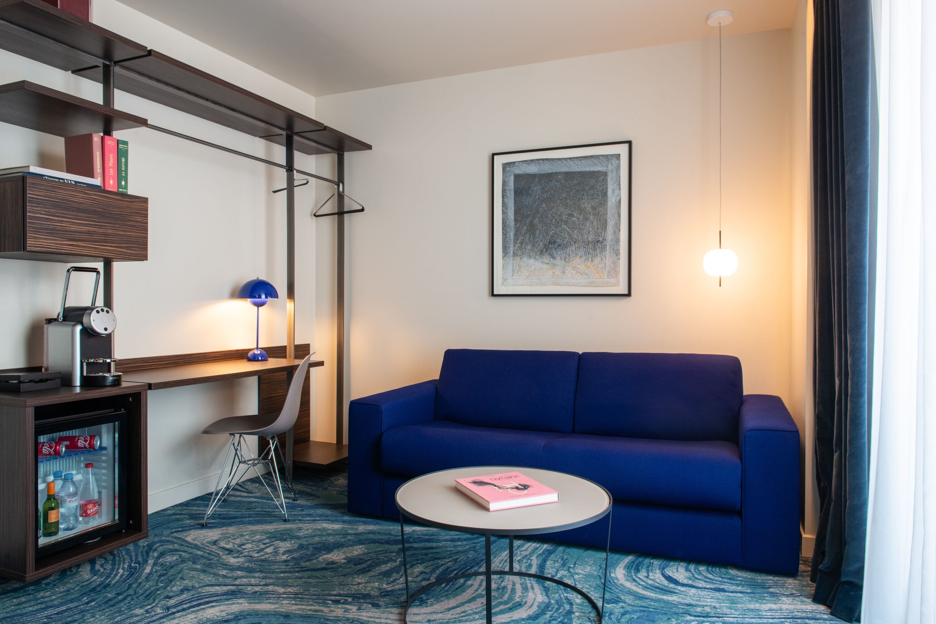 A modern hotel room with a blue sofa, a small round table, a desk with a chair, a coffee machine, and a mini-fridge.