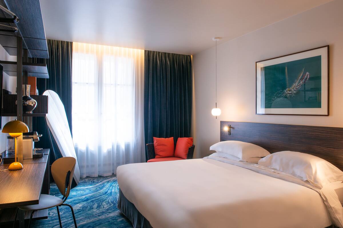 A hotel room with a large bed, a desk with a lamp, a chair, and a window with curtains.