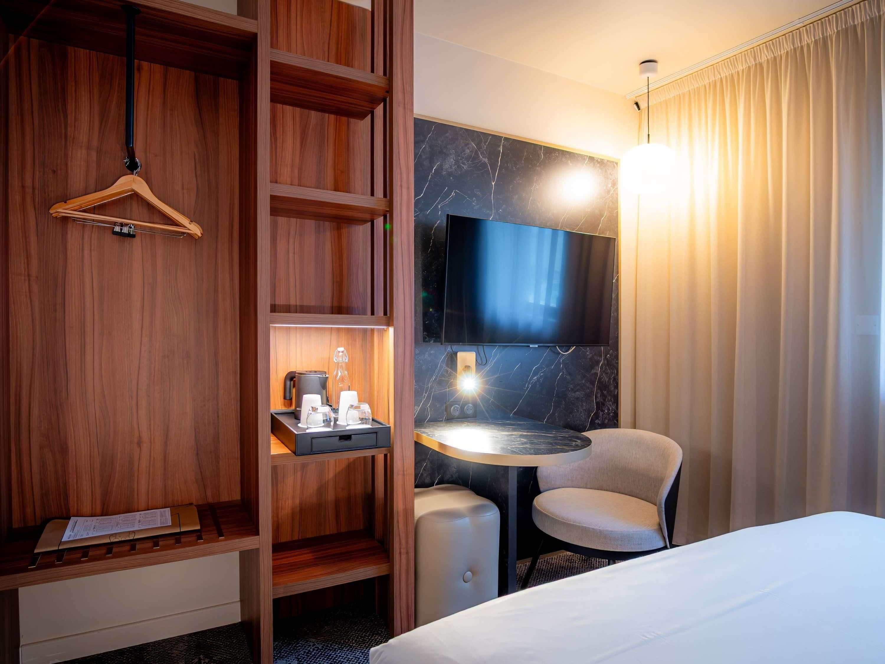 A hotel room with a wooden wardrobe, a flat-screen TV mounted on a marble wall, a small round table with a chair, and a bed with a white bedspread.