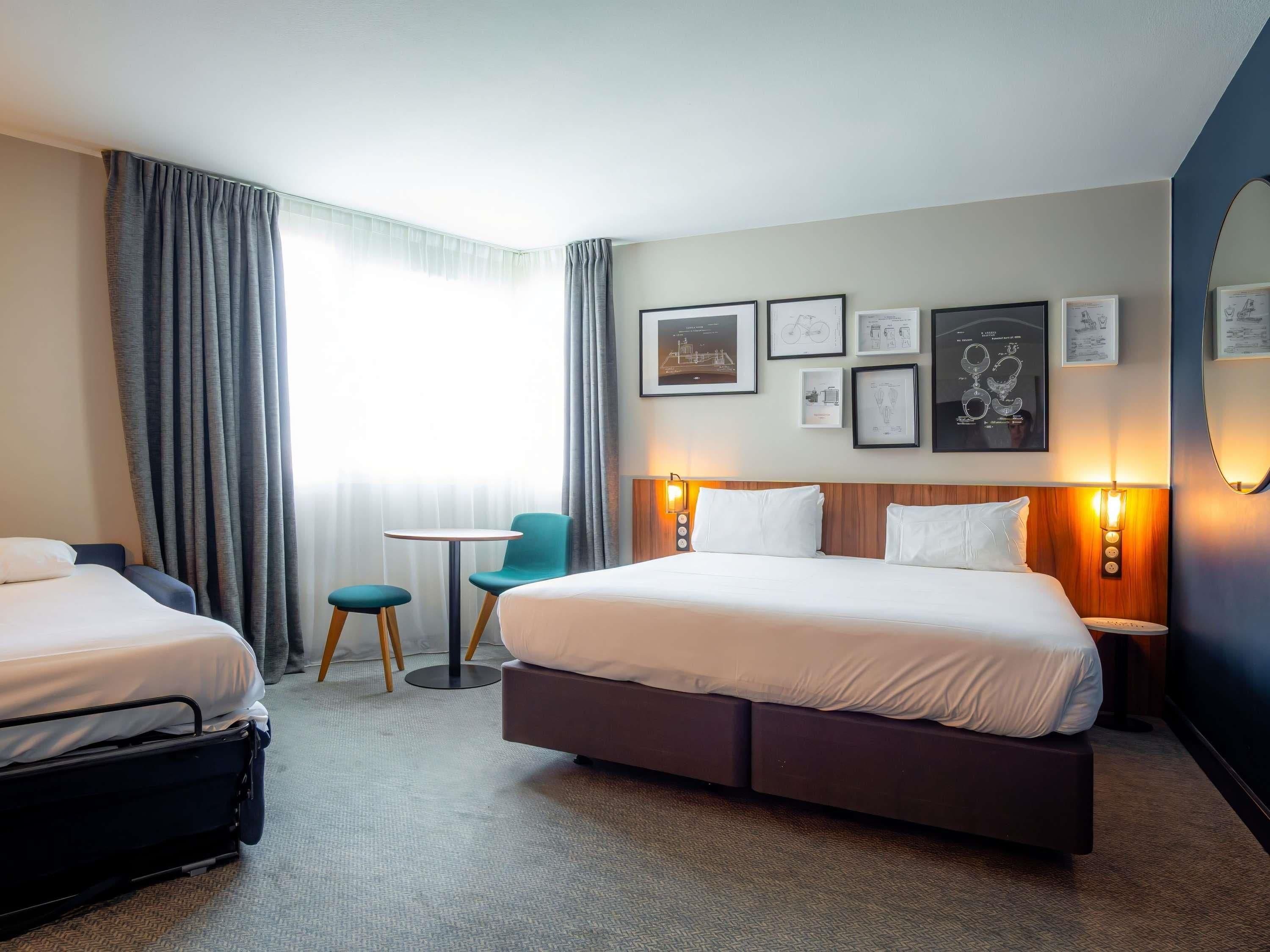 A hotel room with two beds, a small table, chairs, a suitcase, and wall-mounted artwork.