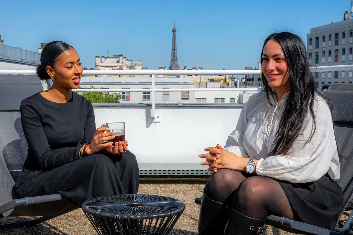 Two people sitting on a rooftop terrace with a view of a cityscape and a tower in the background.