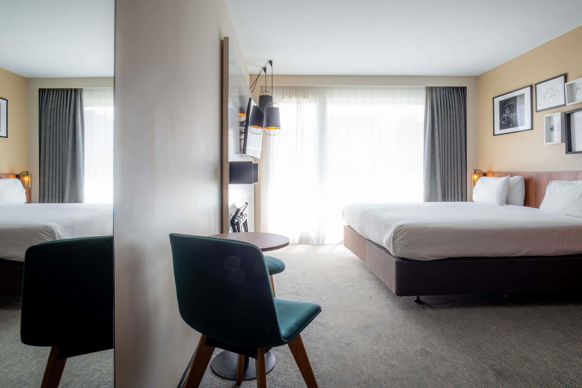 A hotel room with two beds, a desk, a chair, a lamp, and a window with curtains.