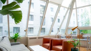 A modern interior with a sofa, ottoman, and a small table, large windows, and a potted plant. - Mercure Paris Montparnasse Pasteur (Paris)
