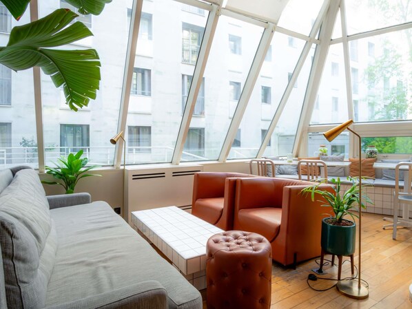 A modern interior with a sofa, ottoman, and a small table, large windows, and a potted plant. - Mercure Paris Montparnasse Pasteur (Paris)