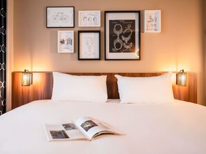 A hotel room with a large bed, two bedside lamps, and framed patent drawings on the wall.