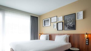 A hotel room with a large bed, a bedside table, a wall-mounted lamp, and framed artwork. - Mercure Paris Montparnasse Pasteur (Paris)