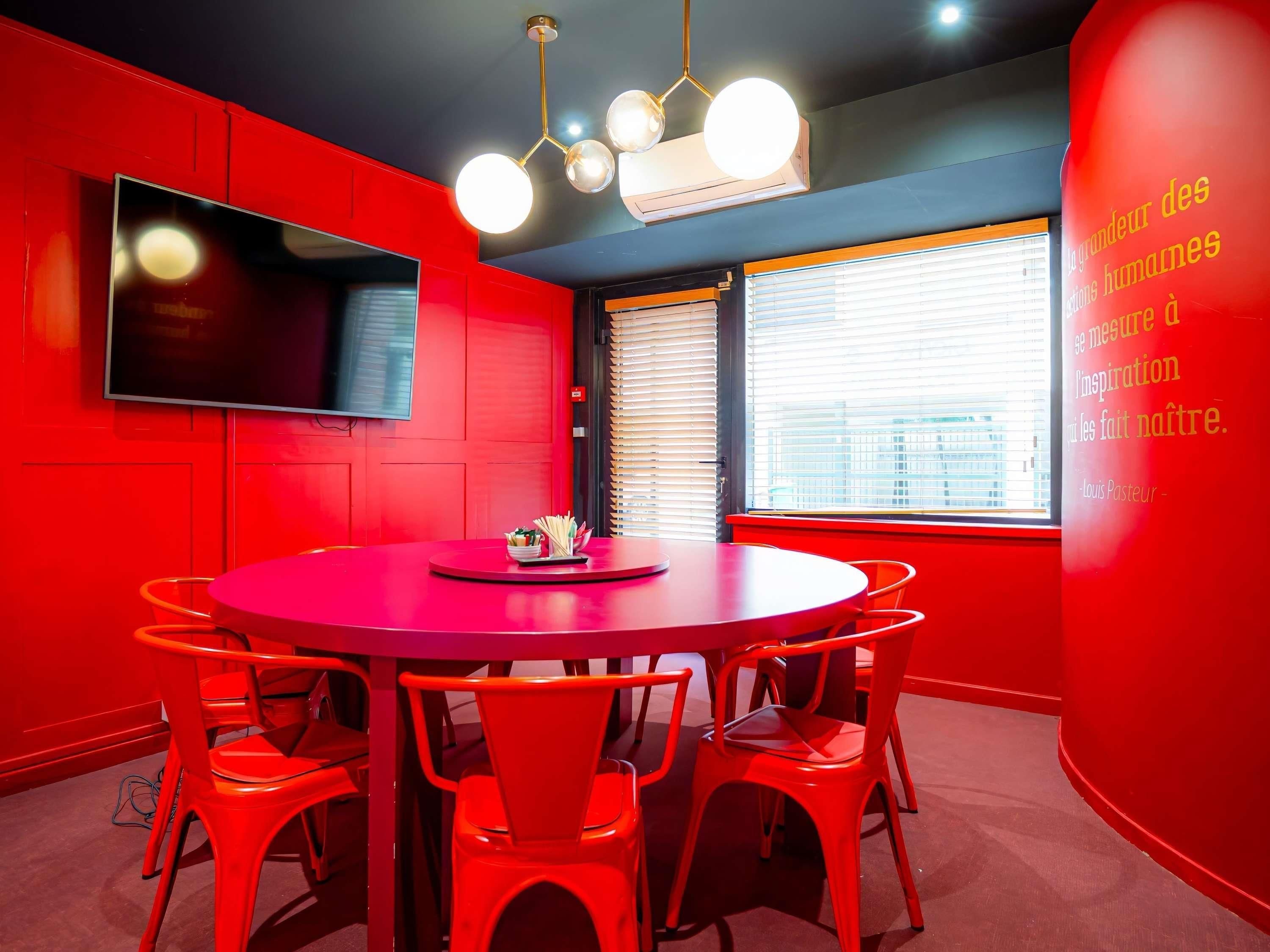 A red-themed meeting room with a round table, chairs, a flat-screen TV, and a large window with blinds.