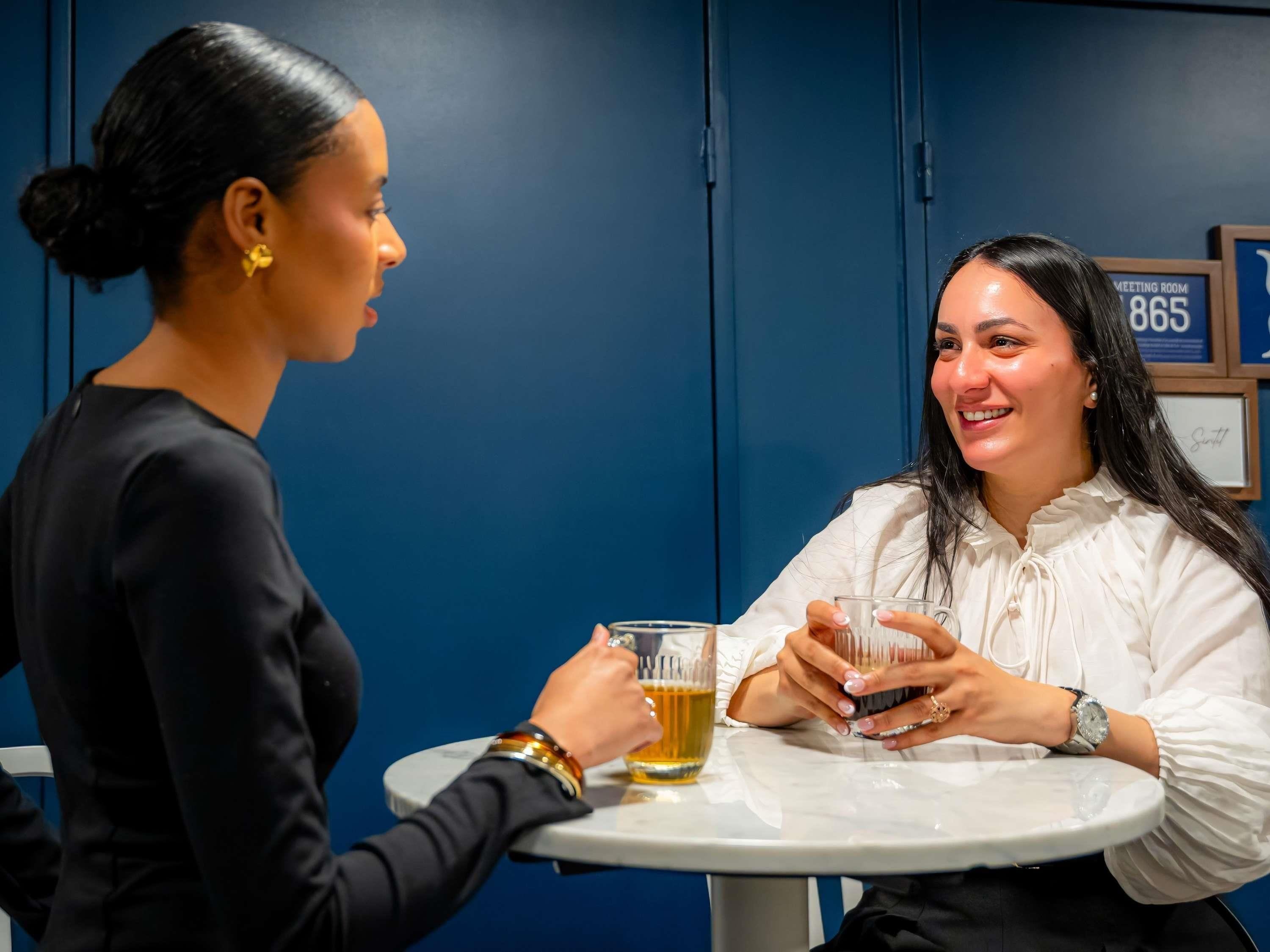 Dos personas sentadas en una mesa redonda en una habitación con paredes azules, sosteniendo vasos de cerveza.